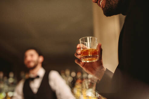 Nahaufnahme eines Mannes an der Bar mit einem Glas Whiskey. Ein Mann nimmt einen Drink an der Bar mit einem Barkeeper im Hintergrund. - JLPPF00088