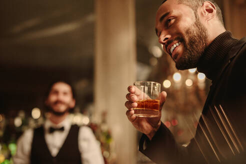 Mann in einem Nachtclub mit einem Glas Whiskey, der wegschaut und lächelt. Mann mit einem Drink an der Bar mit Barkeeper im Hintergrund. - JLPPF00089