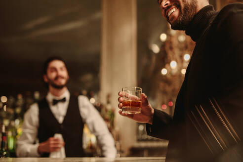 Nahaufnahme eines Mannes, der mit einem Drink an der Bar steht. Ein Mann trinkt einen Whiskey an der Bar mit einem Barkeeper im Hintergrund. - JLPPF00090