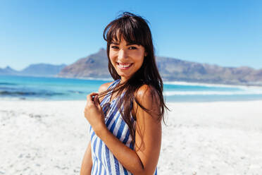 Porträt einer hübschen jungen Frau am Meer. Junges kaukasisches weibliches Modell posiert am Strand, schaut in die Kamera und lächelt. - JLPPF00130
