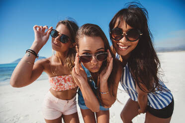 Porträt einer schönen jungen Frau mit Sonnenbrille, die am Strand posiert. Drei Freundinnen stehen zusammen am Meer und schauen in die Kamera. - JLPPF00144
