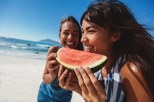 Zwei junge Frauen halten ein Stück Wassermelone und lächeln am Strand. Freundinnen essen Wassermelone am Strand an einem Sommertag. - JLPPF00157
