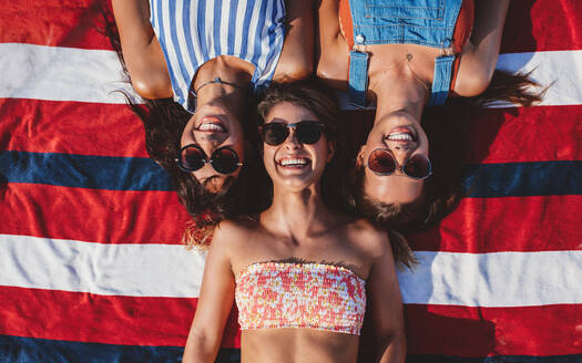 Blick von oben auf drei Freundinnen, die auf einem gestreiften Handtuch am Strand liegen. Junge Frauen am Meer, die den Sommerurlaub genießen. - JLPPF00163