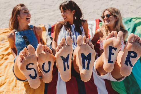 Word summer on the feet of young women sitting at the beach. Female friends at the beach with feet up and enjoying summer holidays. - JLPPF00169