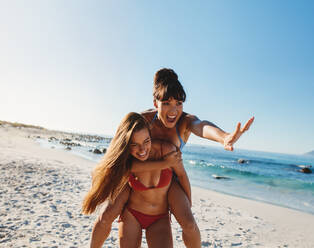 Porträt einer lachenden jungen Frau, die ihre Freundin huckepack nimmt. Zwei Freundinnen spielen am Meer. - JLPPF00199