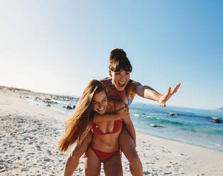 Portrait of laughing young woman giving piggy back ride to her female friend. Two female friends playing at the sea shore. - JLPPF00199