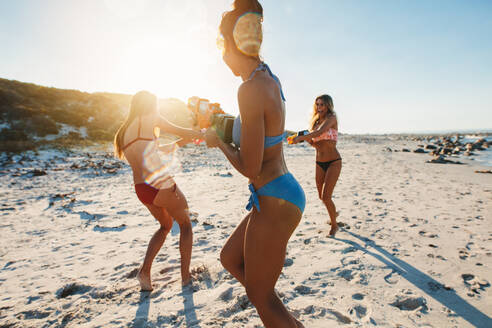 Drei junge Frauen im Bikini liefern sich eine Wasserpistolenschlacht am Meer. Junge Freunde spielen mit Wasserpistolen am Strand. - JLPPF00219