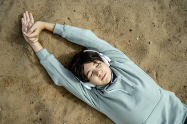 Boy wearing wireless headphones relaxing on sand at beach - ANAF00024