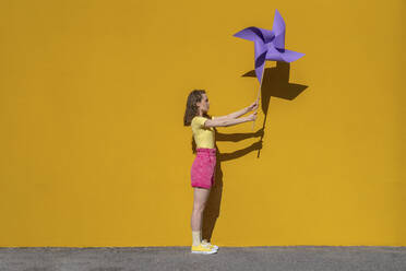 Woman holding pinwheel toy standing in front of vibrant yellow wall - VPIF07306