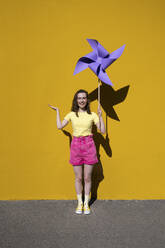 Smiling woman gesturing holding pinwheel toy standing in front of yellow wall - VPIF07314