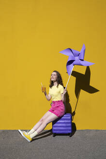Frau mit Getränk und Windrädern auf einem Rollkoffer sitzend vor einer gelben Wand - VPIF07317