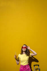 Smiling woman enjoying sunlight holding drink in front of yellow wall - VPIF07323