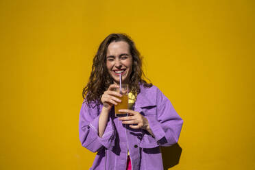 Cheerful woman with eyes closed drinking juice in front of yellow wall - VPIF07328