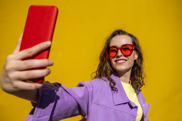Frau mit Sonnenbrille nimmt Selfie in der Nähe von gelben Wand an einem sonnigen Tag - VPIF07343