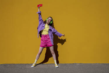 Woman with hand raised enjoying music standing in front of yellow wall - VPIF07352