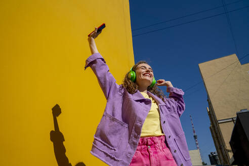 Fröhliche Frau mit erhobener Hand, die an einem sonnigen Tag in der Nähe einer Mauer Musik hört - VPIF07354