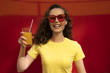 Glückliche Frau mit Sonnenbrille und Getränk vor einer roten Wand - VPIF07356