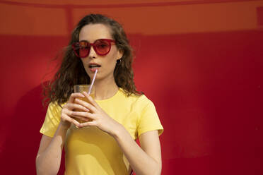 Frau mit Sonnenbrille hält ein Saftglas vor einer roten Wand - VPIF07357