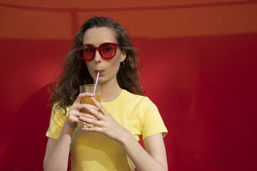 Woman drinking juice wearing sunglasses in front of red wall - VPIF07358