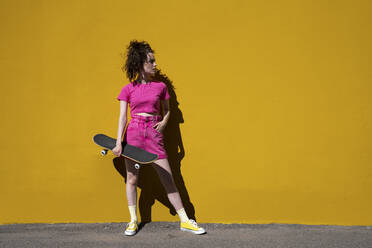 Woman with hand in pocket holding skateboard standing in front of yellow wall - VPIF07367