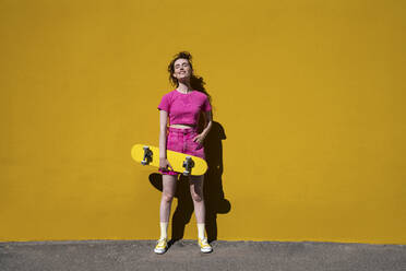 Frau mit Hand in der Tasche, die ein Skateboard hält, steht in der Nähe einer Mauer an einem sonnigen Tag - VPIF07368