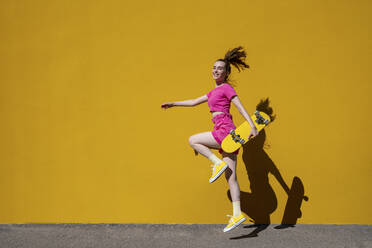 Playful woman with skateboard jumping in front of yellow wall - VPIF07373