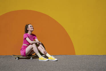 Thoughtful woman sitting on skateboard in front of colorful wall - VPIF07376