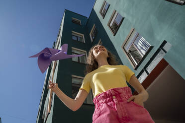 Lächelnde Frau mit Hand in der Tasche, die ein Spielzeug in Form eines Windrades hält, steht in der Nähe eines Gebäudes - VPIF07393