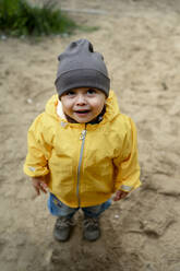 Cute baby boy wearing knit hat and yellow rain jacket on sand - ANAF00032