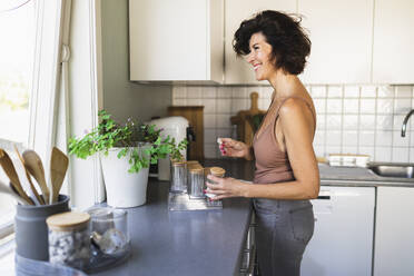 Lächelnde Frau, die ein Glas in der Hand hält und durch ein Fenster schaut, steht am Küchentisch - DMMF00056