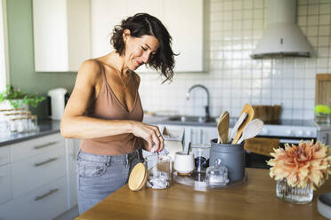 Happy woman filling glass jar at kitchen counter - DMMF00071