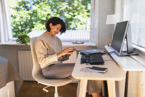 Lächelnde Geschäftsfrau, die ein Smartphone vor einem Desktop-PC auf einem Tisch zu Hause benutzt - DMMF00099