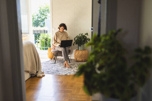 Lächelnde Geschäftsfrau mit Laptop auf einem Stuhl zu Hause sitzend - DMMF00103