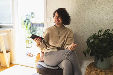 Happy woman holding mobile phone enjoying music with in-ear headphones sitting on chair at home - DMMF00105