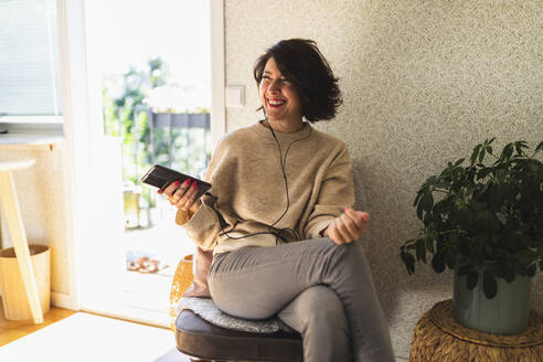 Glückliche Frau mit Handy in der Hand, die mit In-Ear-Kopfhörern auf einem Stuhl sitzend zu Hause Musik hört - DMMF00105