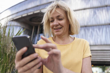 Happy mature woman using smart phone in front of building - TOF00106