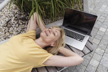 Glückliche blonde Frau mit Laptop auf einer Bank liegend - TOF00118
