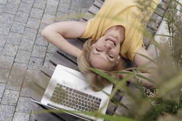 Happy mature woman relaxing by laptop on bench - TOF00119