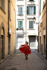 Senior woman in red dress and sun hat dancing on footpath - FLLF00732