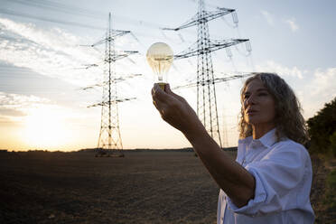 Senior woman looking at light bulb in front of electricity pylons - FLLF00736