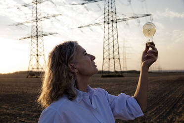 Thoughtful woman looking at light bulb in front of electricity pylons - FLLF00738