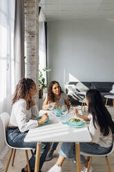 Junge Freunde unterhalten sich und essen Salat am Esstisch im Wohnzimmer - MEUF08047