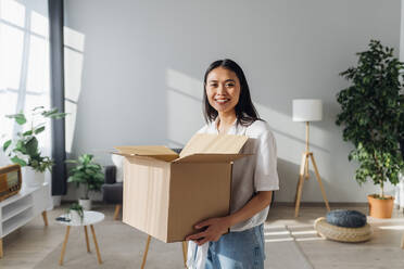 Smiling woman holding cardboard box standing in living room at new home - MEUF08098
