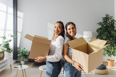 Smiling roommates with cardboard boxes in living room - MEUF08101