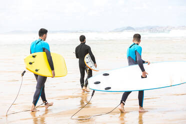 Rückansicht Ganzkörperansicht von fitten jungen ethnischen männlichen Freunden mit dunklen Haaren in Neoprenanzügen, die Surfbretter halten und am Sandstrand spazieren gehen, bevor sie an einem sonnigen Tag surfen - ADSF38684