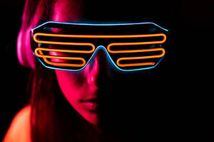 Junge Frau mit futuristischer Brille hört Musik mit drahtlosen Kopfhörern unter rotem Neonlicht in dunkler Nacht - ADSF38698