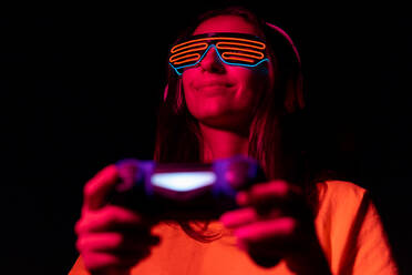 From below happy young woman in futuristic glasses smiling and using gamepad while playing videogame under red neon light at night - ADSF38699