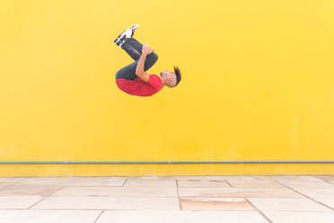 Male jumping near yellow wall and showing somersault while doing parkour in city on sunny day - ADSF38700