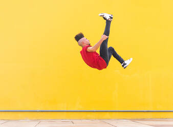 Male jumping near yellow wall and showing somersault while doing parkour in city on sunny day - ADSF38701