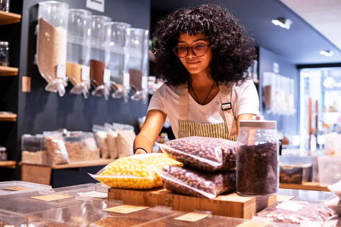 Positive junge schwarze Frau in Uniform und mit Brille, die lächelt und Plastiktüten mit Puffcorn-Snacks in ein Regal neben einem Glas mit Teeblättern stellt, während sie in einem Zero-Waste-Lebensmittelladen arbeitet - ADSF38705